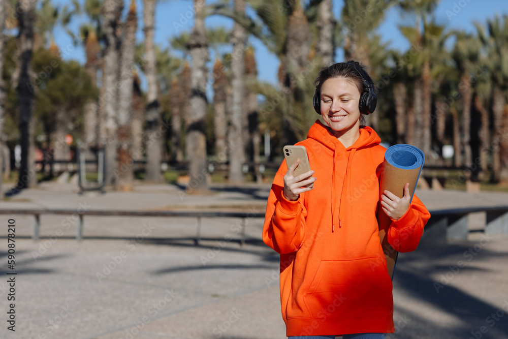 Joyous young woman In headphones with yoga mat, listens music on smartphone outdoors. Happy millennial lady in vivid orange sweatshirt enjoying favorite playlist while has break in training or after