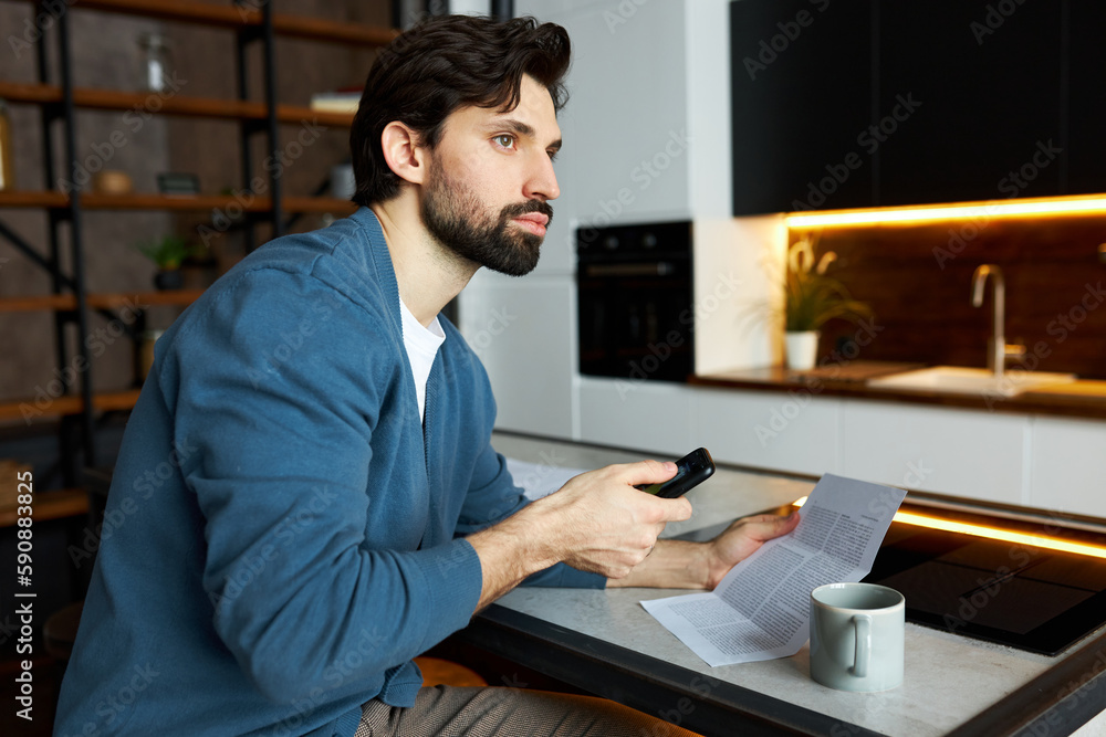 Side view portrait of handsome businessman reading paper document holding mobile phone in hands, dialing number of his assistant, sitting at kitchen counter, looking aside with thoughtful face