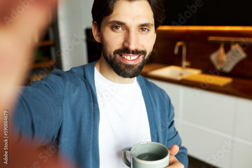 Cheerful positive bearded guy blogger drinking hot water and recording stories for social media news feed, smiling widely standing with cup against kitchen sink. People and technology