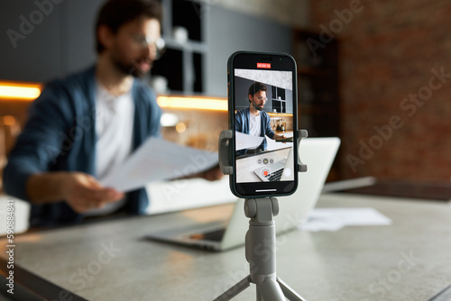 Selective focus on tripod with smartphone recording male business coach sitting at kitchen table in front of laptop, holding paper documents, preparing for live stream. Vlogging concept