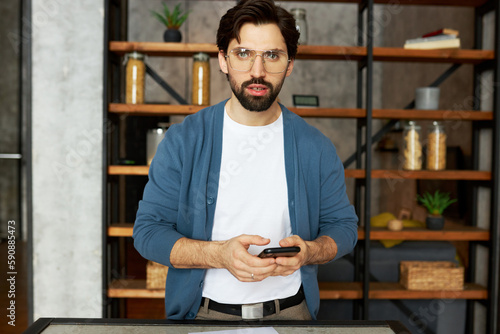 Serious thoughtful office worker standing with smartphone in hands, trying to remember number and make important urgent call, posing against loft style interior in glasses and blue cardigan