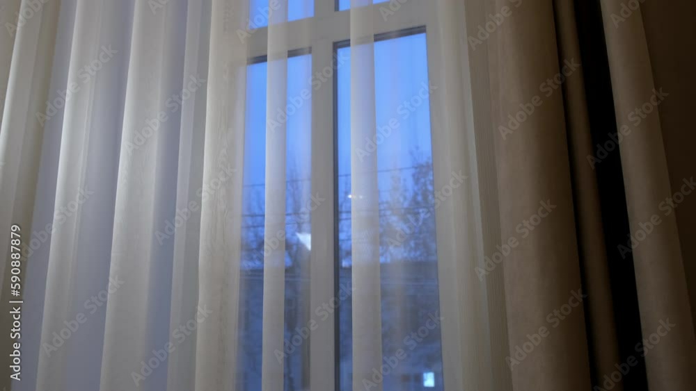 Beige curtaing on the window with fine tulle in modern interior in minimalism style. Winter evening city outside the window. Beige curtaing on the window with fine tulle in modern interior in minimalism style. Winter evening city outside the window.