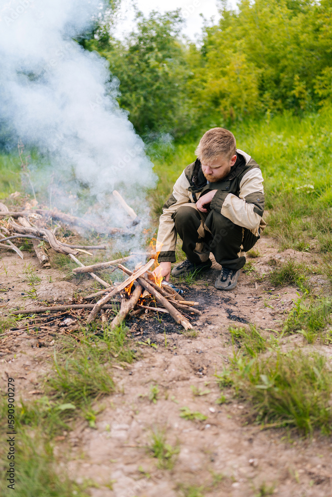 Vertical portrait of lost survivalist male in raincoat putting firewood on campfire to making fire on bank of river in evening before sunset. Concept of exploration, travel and adventure.