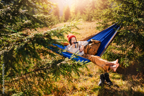 Teenage child lies in travel hammock and drinks tea from metal travel mug. Recreation in the fresh air concept.