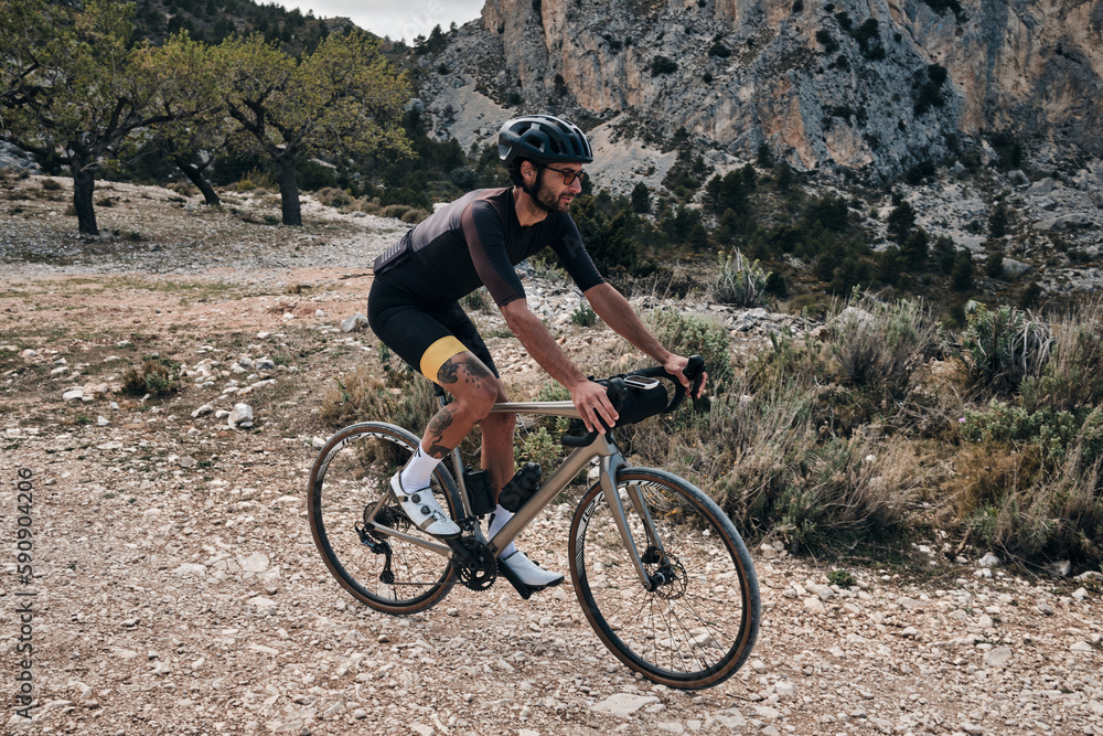 Naklejka premium Man riding gravel bike on gravel road in mountains with scenic view.Professional cyclist practicing on gravel road.Male cyclist wearing black cycling kit and helmet.Cycle camp in Calpe, Alicante,Spain
