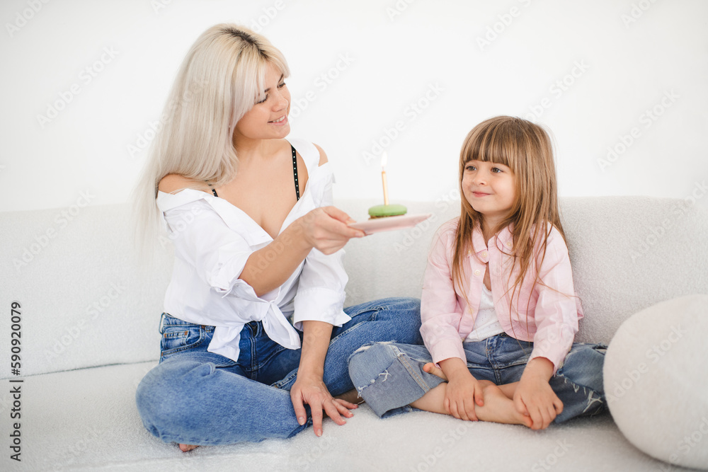 Cute funny kid girl 4-5 year old with young mother holding birthday biscuit with candle sitting on cozy sofa in room over white at home. Motherhood. Cute funny kid girl 4-5 year old with young mother holding birthday biscuit with candle sitting on cozy sofa in room over white at home. Motherhood.