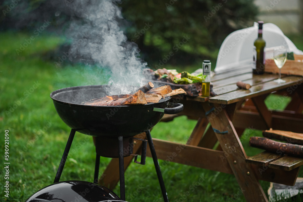 The smoking brazier of a round form. Firewood flares up in the brazier against the background of a table and chairs. The concept of picnic, relaxation, grilling. The smoking brazier of a round form. Firewood flares up in the brazier against the background of a table and chairs. The concept of picnic, relaxation, grilling.