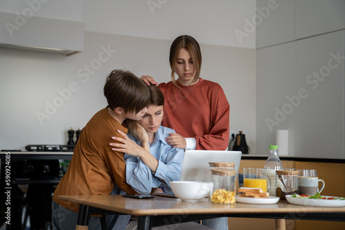 Loving children supporting hugging single mother struggling to find job. Upset unhappy family mom and two kids embracing looking at laptop screen. Son and daughter support upset mum lost job