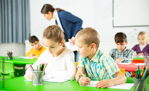 Portrait of diligent preteen students writing exercises at lesson with young female teacher in primary school