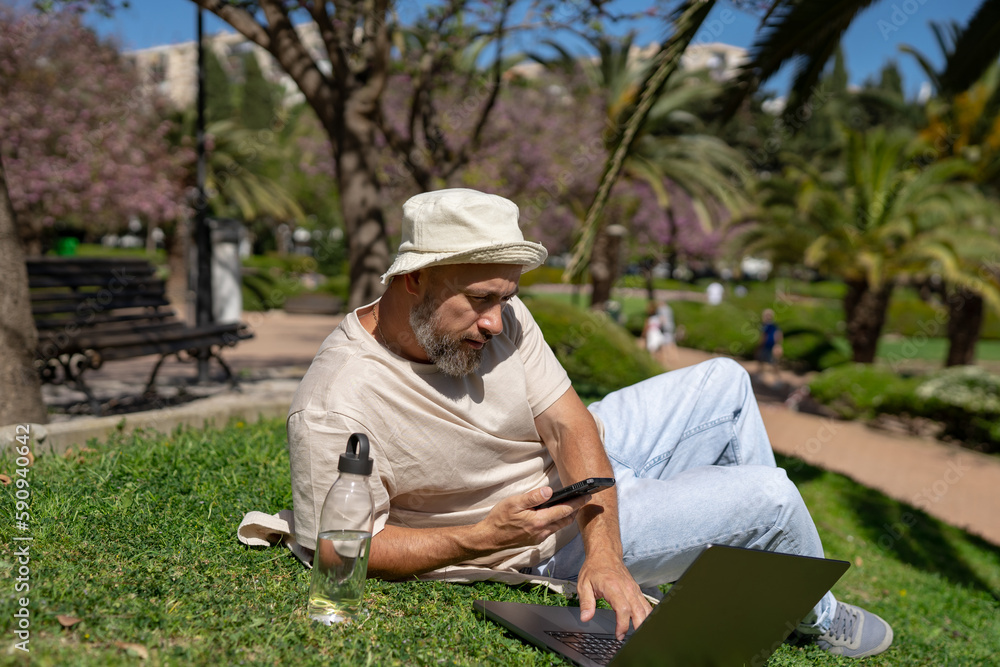 bearded male freelancer with a mobile phone in his hands works on a laptop while sitting on green grass in a park on a sunny summer day