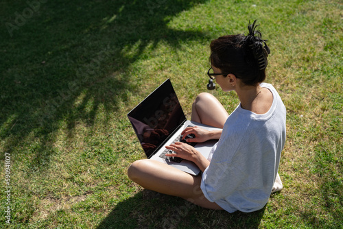 a beautiful girl freelancer in glasses works at a laptop in the summer in the park against the backdrop of palm trees next to a bottle of water