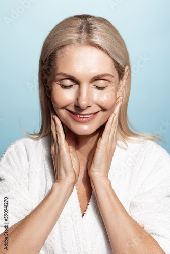 Aging beauty. Vertical portrait of middle aged, elderly woman, 50 years, touches her smooth, nourished skin, applies facial cream, moisturizer, smiling pleased, wears bathrobe, blue background