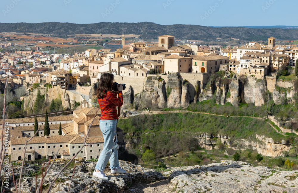 Young female tourist with a camera, climbing on top of a cliff, photographs a picturesque landscape with a view of the city ..of Cuenca, Spain