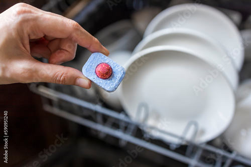 Close up of woman hand filling dishwasher tablet into open automatic stainless built-in dishwasher machine with dirty white dish inside in modern home kitchen.Household, housekeeping domestic life. photo