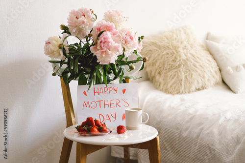 Breakfast for Mothers Day. Heart shaped white plate with fresh strawberries, cup of coffee, gift and Peonys bouquet with gift in bed. Still life composition. Happy Mother's Day.