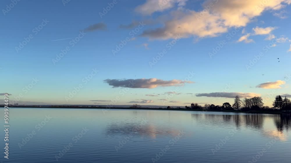 Blue lake water with golden sunset sky, dreamy clouds. Perfect winter evening. Pastel blue shade mirroring monochrome sky over water surface. Wild birds flying above below distance. Boat perspective Blue lake water with golden sunset sky, dreamy clouds. Perfect winter evening. Pastel blue shade mirroring monochrome sky over water surface. Wild birds flying above below distance. Boat perspective
