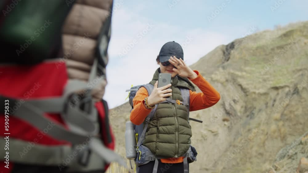 Medium shot of young Asian female tourist making video of her friend, partly seen from behind, standing against background of mountain. Daytime, cold weather Medium shot of young Asian female tourist making video of her friend, partly seen from behind, standing against background of mountain. Daytime, cold weather