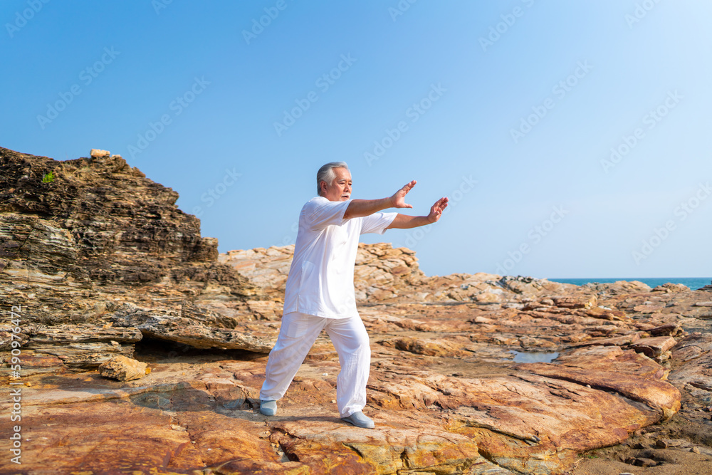 Asian senior man practicing meditation with nature on rocky coastal hill. Retirement elderly people do outdoor relaxing tai chi yoga exercise at summer sunset. Mental health care motivation concept. Asian senior man practicing meditation with nature on rocky coastal hill. Retirement elderly people do outdoor relaxing tai chi yoga exercise at summer sunset. Mental health care motivation concept.