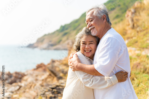 Happy Asian family senior couple with gray hair relaxing and hugging each other on mountain cliff at sunset. Retirement elderly people enjoy outdoor lifestyle travel nature ocean on summer vacation.