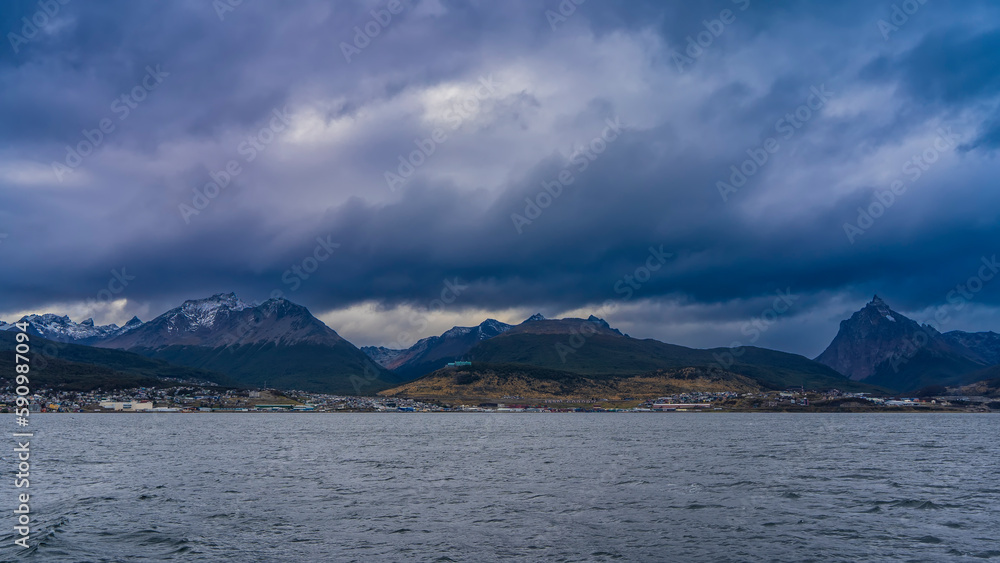 A picturesque snow-capped mountain range against the background of the sky and dark rain clouds. City houses on the shore of the strait. Beagle Channel. Argentina. Ushuaia