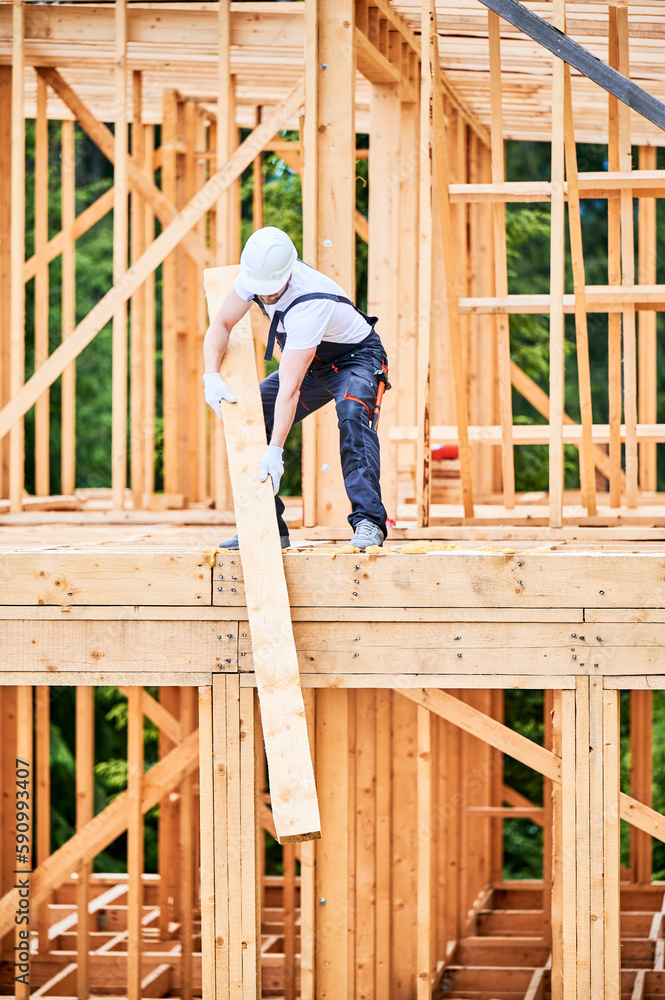Carpenter building wooden-framed house. Man lifting up large beam, dressed in work clothes and helmet. Concept of contemporary and eco-friendly construction. Carpenter building wooden-framed house. Man lifting up large beam, dressed in work clothes and helmet. Concept of contemporary and eco-friendly construction.