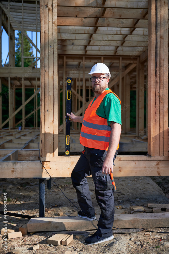 Carpenter building wooden frame two-story house. Bearded man in glasses holding a level, wearing protective helmet, overalls and orange vest. Concept of modern ecological construction. Carpenter building wooden frame two-story house. Bearded man in glasses holding a level, wearing protective helmet, overalls and orange vest. Concept of modern ecological construction.