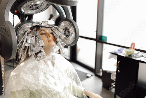 Φωτογραφία Portrait of young woman sitting in salon with hair dye applied on long hair wrapped in foil under hooded dryer machine