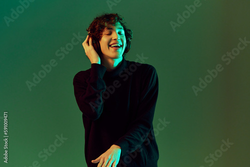 Man wearing headphones listening to music, dancing and singing with his eyes closed, DJ happiness and smile laughter, hipster teen lifestyle, portrait green background mixed neon light, copy space © SHOTPRIME STUDIO