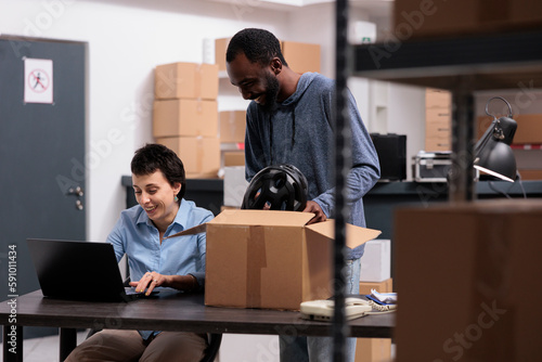 African american worker putting client helmet order in carton box preparing delivery for customer while discussing transportation details with manager. Diverse team working in warehouse