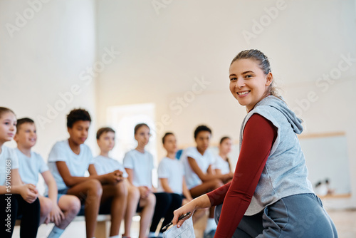 Young happy sports teacher having PE class with group of kids at school gym and looking at camera.