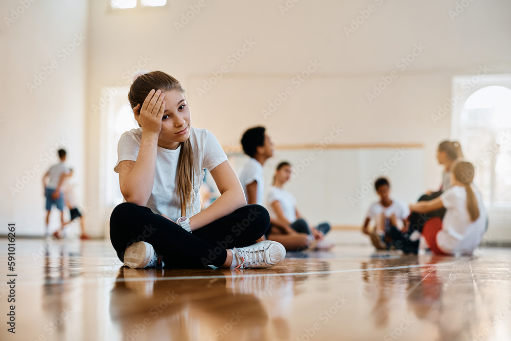 Sad elementary student sits away from her classmates and teacher during PE class at school. Sad elementary student sits away from her classmates and teacher during PE class at school.