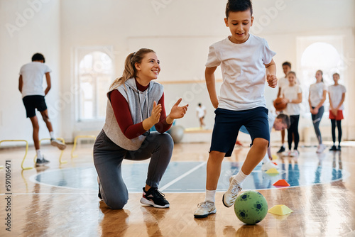 Schoolboy leads soccer ball among cones while coach is supporting him during physical activity class.