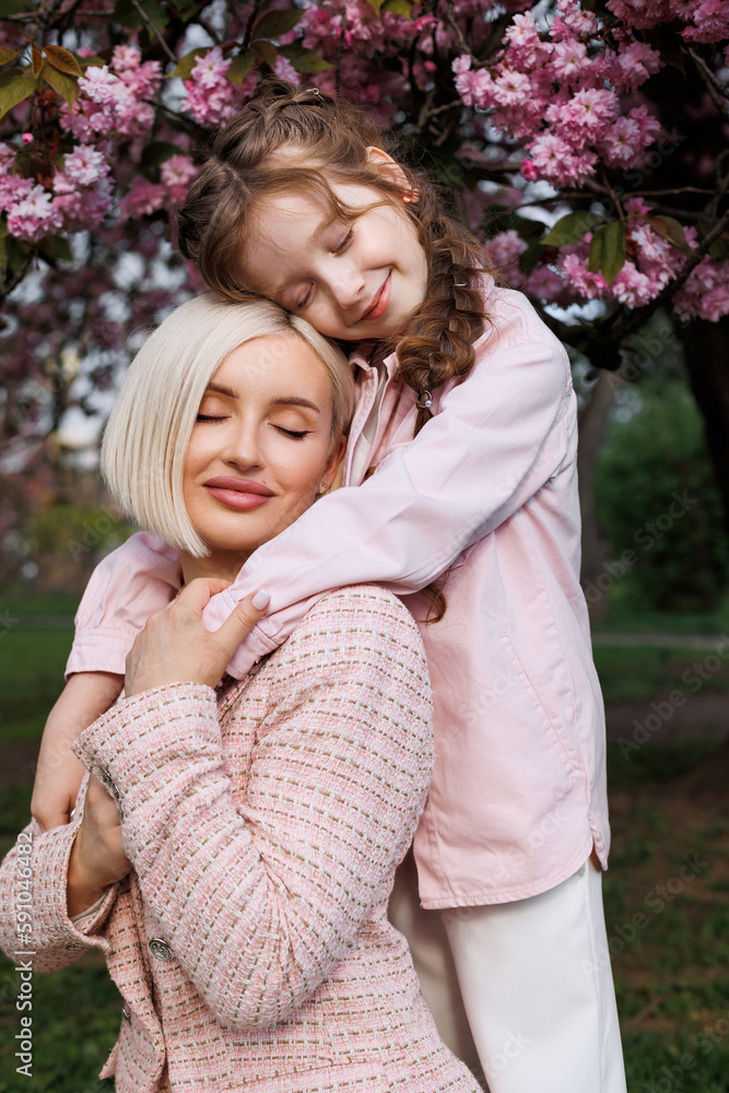 Fototapeta premium A daughter stands on top and hugs her happy mother tightly under a cherry tree on a sunny spring day