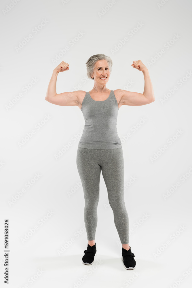 full length of joyful and retired woman in sportswear showing muscles on grey. full length of joyful and retired woman in sportswear showing muscles on grey.