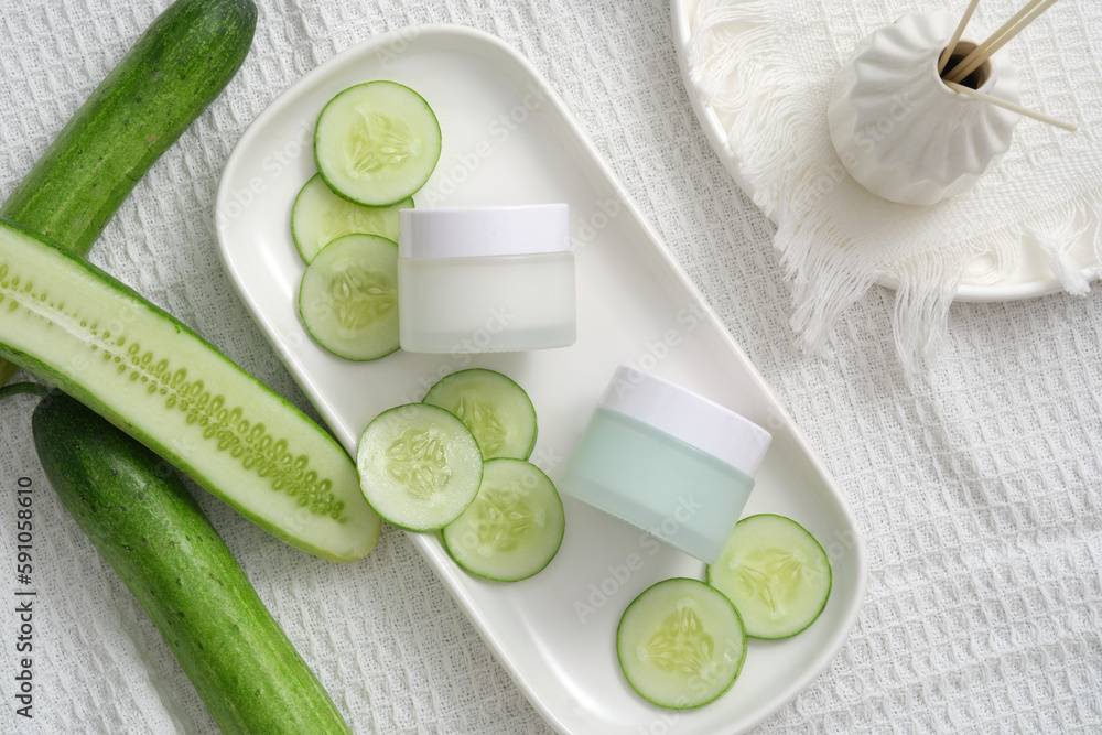 Cream jars unlabeled and cucumber slices on white fabric background. Mockup for cosmetic of cucumber extract. The high water content and antioxidants in cucumber make the skin supple and elastic. Cream jars unlabeled and cucumber slices on white fabric background. Mockup for cosmetic of cucumber extract. The high water content and antioxidants in cucumber make the skin supple and elastic.
