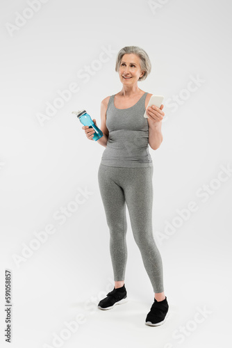 full length of happy senior woman in sportswear holding sports bottle with water and smartphone on grey.