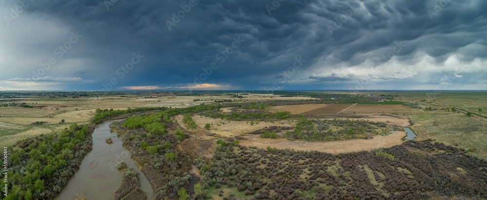 Aerial panoramic shot of a field with a narrow lake on it on a cloudy day