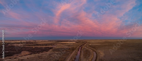 Fototapeta Naklejka Na Ścianę i Meble -  Aerial panoramic shot of a field with a narrow lake on it during a pink sunset