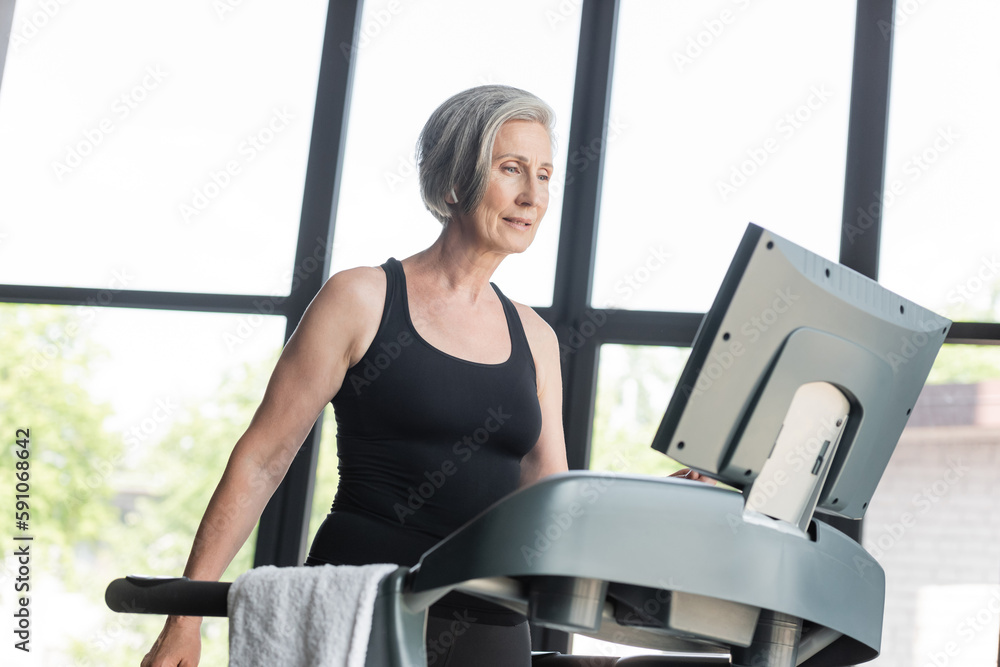 retired woman with grey hair looking at monitor of treadmill while working out in gym. retired woman with grey hair looking at monitor of treadmill while working out in gym.