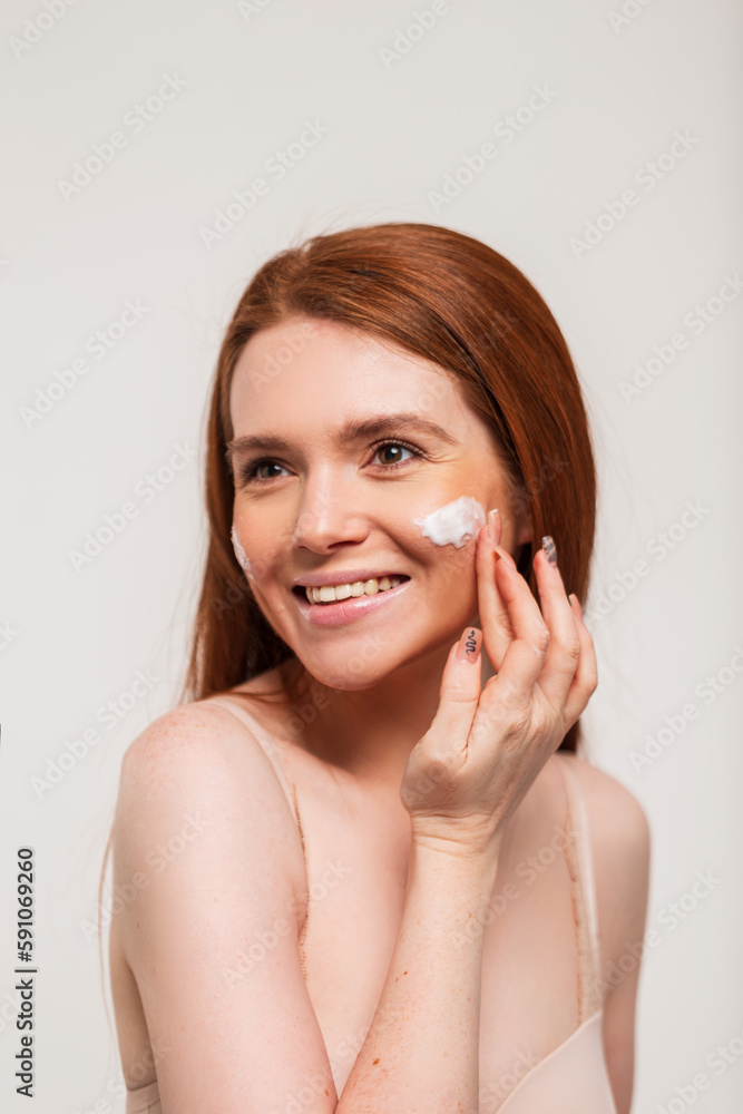 Happy beautiful young redhead woman with a cute smile in a beige bra applies cream on her face skin on a white background in the studio. Beauty and skin care Happy beautiful young redhead woman with a cute smile in a beige bra applies cream on her face skin on a white background in the studio. Beauty and skin care