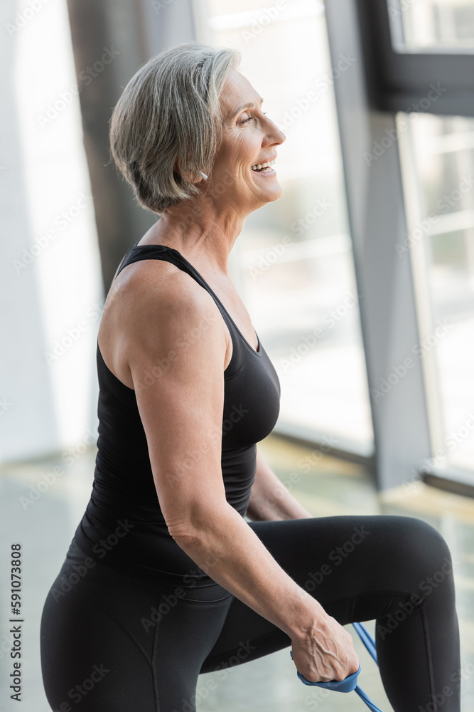 side view of happy senior sportswoman in black leggings and tank top exercising with resistance band.
