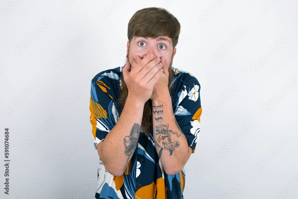 Upset red haired man wearing printed shirt over white studio background, covering her mouth with both palms to prevent screaming sound, after seeing or hearing something bad.