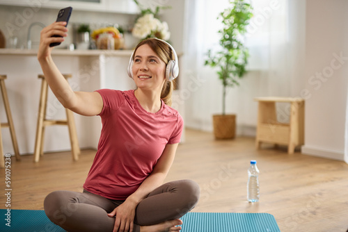 Fototapeta Naklejka Na Ścianę i Meble -  Young woman using cellphone and taking Selfie while exercising yoga at home