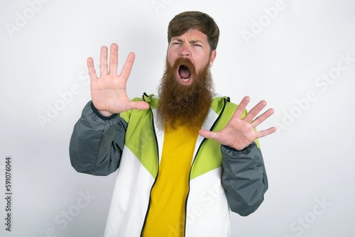 Dissatisfied red haired man wearing sportswear standing over white studio background frowns face, has disgusting expression, shows tongue, expresses non compliance, irritated with somebody. photo