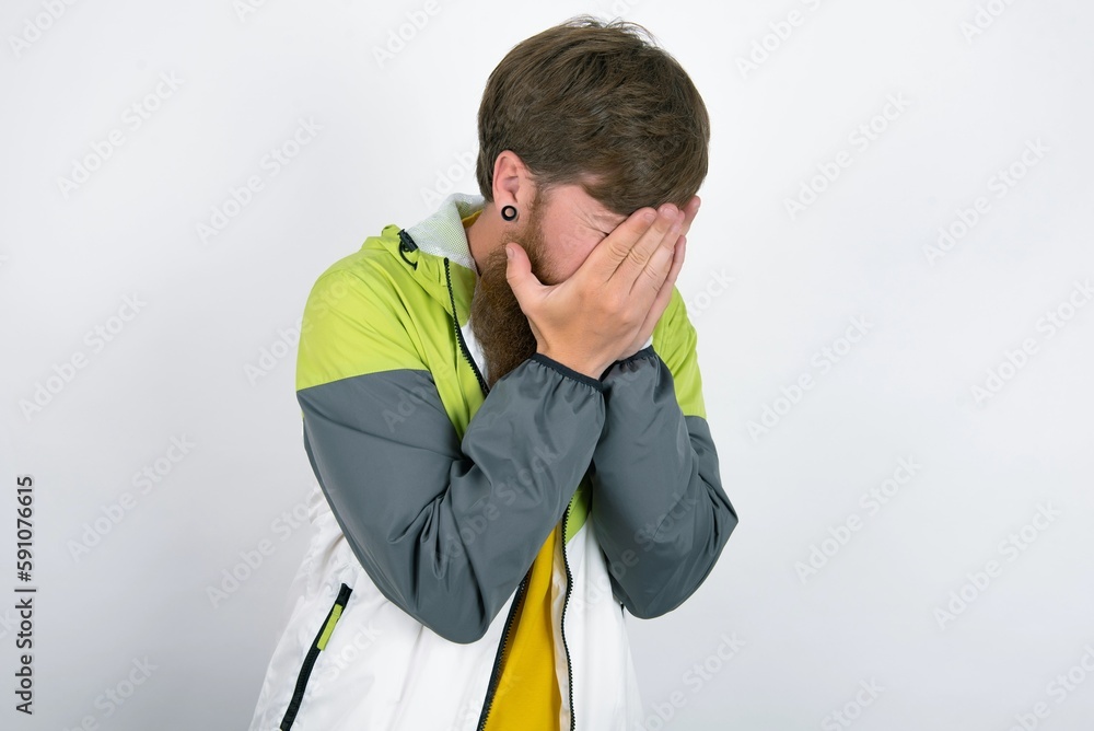 Sad red haired man wearing sportswear standing over white studio background covering face with hands and crying. Sad red haired man wearing sportswear standing over white studio background covering face with hands and crying.