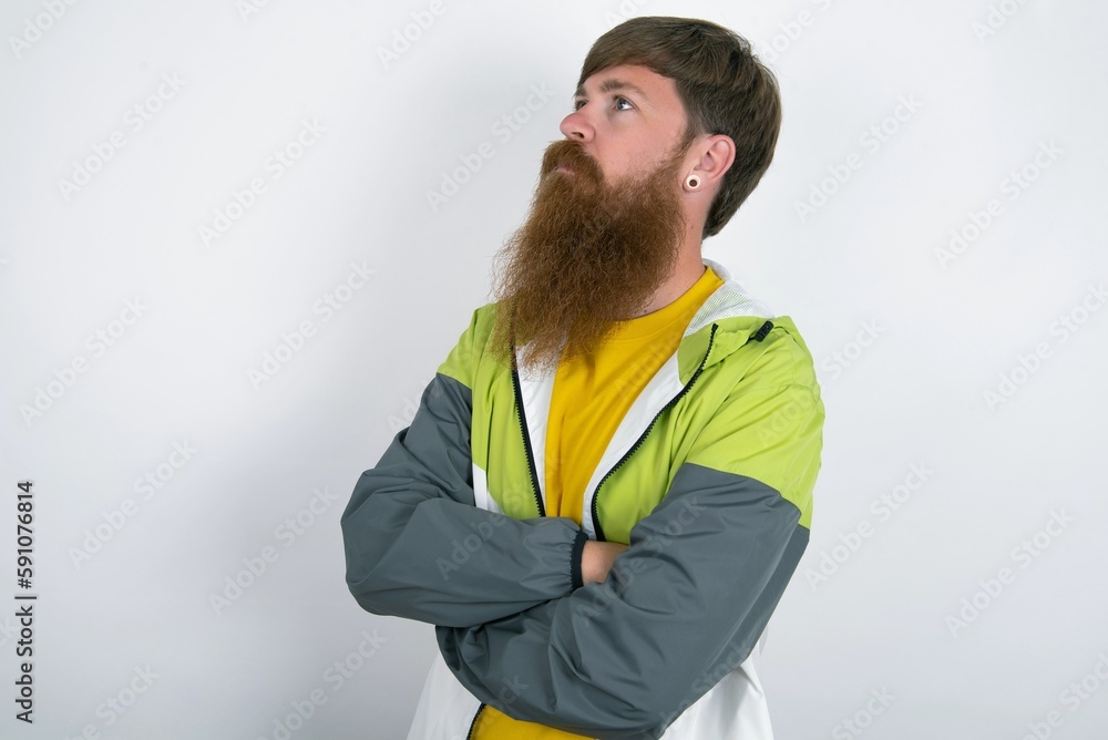 Charming thoughtful red haired man wearing sportswear standing over white studio background stands with arms folded concentrated somewhere with pensive expression thinks what to do Charming thoughtful red haired man wearing sportswear standing over white studio background stands with arms folded concentrated somewhere with pensive expression thinks what to do