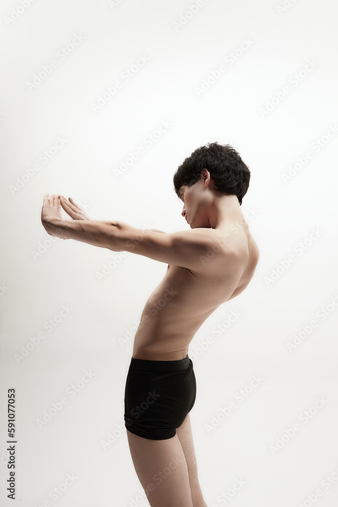 Stretching. Studio image of young guy, man standing shirtless in underwear against white background. Slim body shape. Concept of male body aesthetics, style, fashion, health, men's beauty