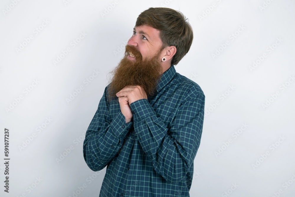 Happy red haired man wearing plaid shirt over white studio background anticipates something awesome happen, looks happily aside, keeps hands together near face, has glad expression. Happy red haired man wearing plaid shirt over white studio background anticipates something awesome happen, looks happily aside, keeps hands together near face, has glad expression.