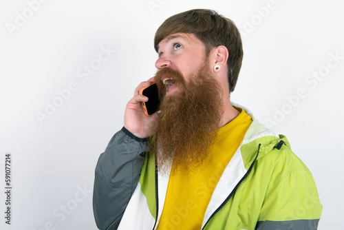 red haired man wearing sportswear standing over white studio background speaks on mobile phone spends free time indoors calls to friend.