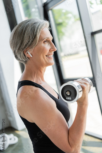 side view of cheerful senior sportswoman in wireless earphone exercising with dumbbell in gym.